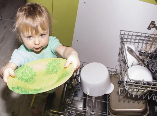 Imádnivaló mosolygós szőke totyogó fiú segít a konyhában, figyelembe véve a lemezek a mosógép — Stock Fotó