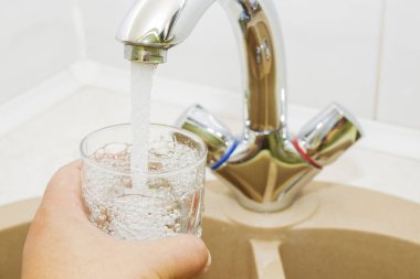 Mutfak musluğundan dökülen bir bardak suyu tutan el.