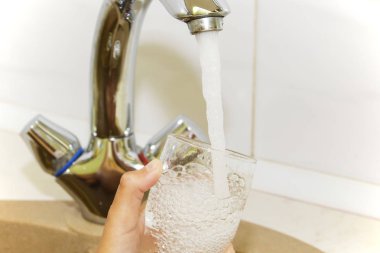 Mutfak musluğundan dökülen bir bardak suyu tutan el.