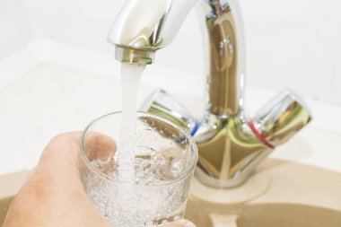 Mutfak musluğundan dökülen bir bardak suyu tutan el.