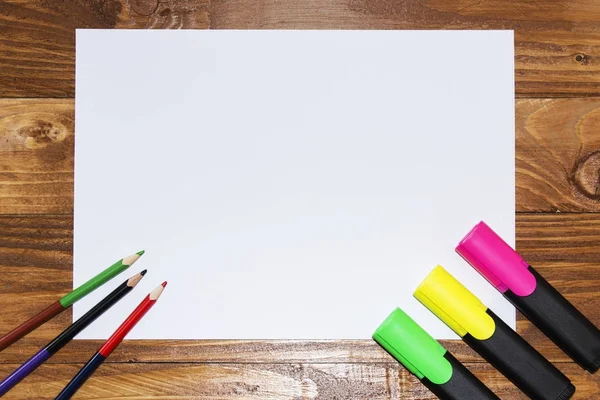 Una hoja blanca de papel yacía sobre una mesa de madera, cerca, lápices, marcadores, bolígrafos. La vista desde arriba . — Foto de Stock