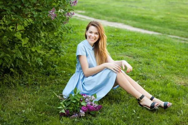 Joven mujer hermosa con el pelo rojo se sienta en una hierba bajo el árbol lila y disfruta de su vida — Foto de Stock