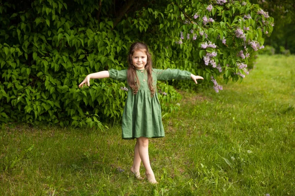 Cute girl in green linen dress has fun in the park with blooming lilacs, enjoys spring and warmth. Beautiful spring garden. Happy childhood, peace and happiness concept