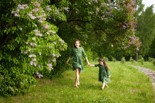 Two sisters in green linen dress have fun in the park with blooming lilacs, enjoy spring and warmth. Beautiful spring garden. Happy childhood. Family, love, peace and happiness concept