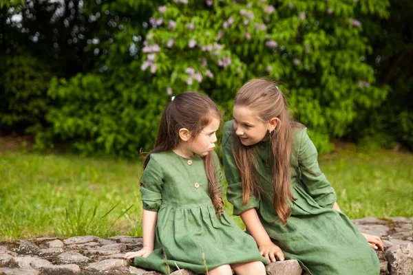 Two sisters in green linen dress have fun in the park with blooming lilacs, enjoy spring and warmth. Beautiful spring garden. Happy childhood. Family, love, peace and happiness concept