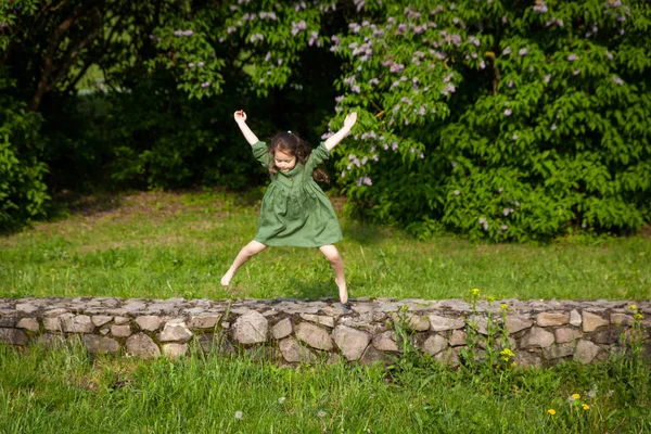 Cute girl in green linen dress has fun in the park with blooming lilacs, enjoys spring and warmth. Beautiful spring garden. Happy childhood, peace and happiness concept