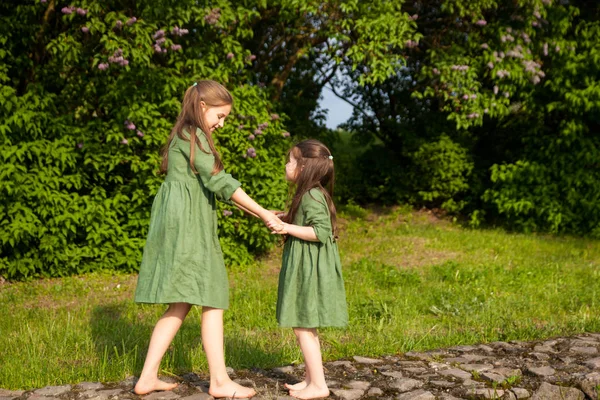 Two sisters in green linen dress have fun in the park with blooming lilacs, enjoy spring and warmth. Beautiful spring garden. Happy childhood. Family, love, peace and happiness concept