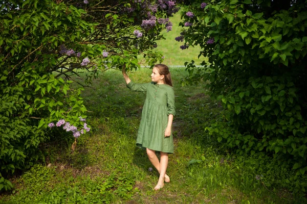 Cute girl in green linen dress has fun in the park with blooming lilacs, enjoys spring and warmth. Beautiful spring garden. Happy childhood, peace and happiness concept