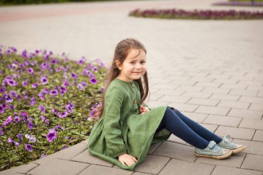 Keten yeşili elbiseli gülümseyen kız mor petunyalı bir çiçek tarlasının yanında oturuyor.