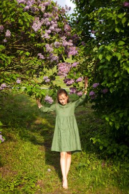 Yeşil keten elbiseli tatlı kız parkta çiçek açan leylaklarla eğleniyor, bahar ve sıcağı seviyor. Güzel bir bahar bahçesi. Mutlu çocukluk, barış ve mutluluk kavramı