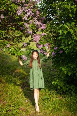Yeşil keten elbiseli tatlı kız parkta çiçek açan leylaklarla eğleniyor, bahar ve sıcağı seviyor. Güzel bir bahar bahçesi. Mutlu çocukluk, barış ve mutluluk kavramı