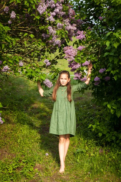 Cute girl in green linen dress has fun in the park with blooming lilacs, enjoys spring and warmth. Beautiful spring garden. Happy childhood, peace and happiness concept