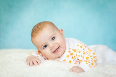 Mutlu 4 ay boyunca beyaz kürklü bir bebek yatakta yattı. Parlak mavi arka planda turuncu benekli beyaz elbiseler giyen tatlı bir kız. Çocuklar için tekstil ve yatak
