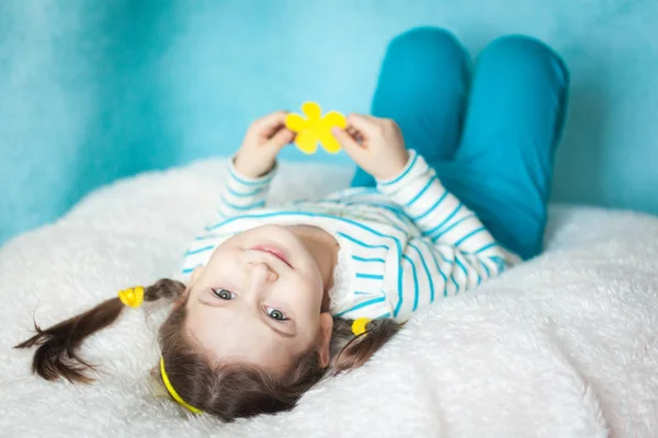 Cute portrait of a girl with pigtails upside down, lying on bed, smiling at camera and playing with paper flower. Blue background and clothes and yellow details. Contrast colors concept. DIY flower gift for mother