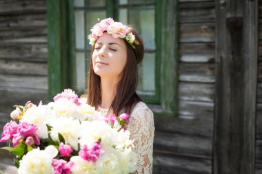 Güzel genç bir kadın, eski ahşap perdeli yeşil pencereli evin yanında büyük şakayık desenli metal bir kova tutuyor.