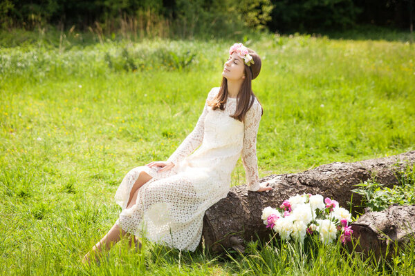 Beautiful young woman in a floral wreath sitting on a big old log in summer garden
