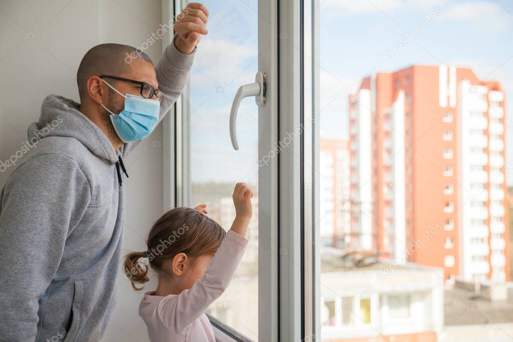 Little girl and her father in medical mask looking through the window. Isolation at home for self quarantine. Concept home quarantine, prevention COVID-19. Coronavirus outbreak situation
