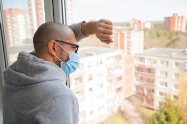 Camdan içeri bakan maskeli bir adam. Kendini karantinaya almak için evde tecrit. Ev karantinası, COVID-19 'u önleme. Coronavirüs salgını durumu