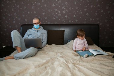 Korunaklı maskeli baba karantina sırasında Coronavirus 'ta çalışıyor. Çocuk evde, anaokulu kapalı. Kızı kitabı okuyor..