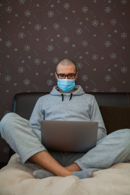 Korunaklı maskeli bir adam Coronavirus Covid-19 salgınında karantina sırasında evden çalışıyor. Dizüstü bilgisayarla yatakta oturuyor. Ev kavramından çalış.