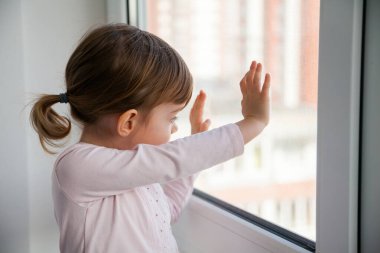 Küçük kız pencereden dışarı bakıyor. Yürümek ve oynamak istiyor ama kendini karantinaya almak için evde kalmalı. Ev karantinası, COVID-19 önleme. 