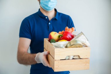Koruyucu tıbbi maskeli ve eldivenli kurye yiyecek dolu bir kutu tutuyor. Çevrimiçi gıda siparişi, karantina sırasında teslimat, koronavirüs salgını. Servis karantina pandemik koronavirüsü.
