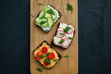 Süzme peynir, humus, avokado, turp ve domatesli sağlıklı vejetaryen çavdar ekmeği. Siyah zemin üzerinde tahta tahta tahta, düz yatıyordu.