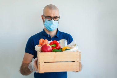 Koruyucu tıbbi maskeli ve eldivenli kurye elinde farklı yiyeceklerle dolu bir kutu tutuyor. Servis karantina pandemik koronavirüsü. Çevrimiçi gıda siparişi, karantina sırasında teslimat, koronavirüs salgını..