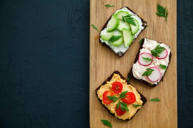 Süzme peynir, humus, avokado, turp ve domatesli sağlıklı vejetaryen çavdar ekmeği. Siyah arkaplan üzerinde tahta tahta tahta, kopyalama alanı, üst görünüm