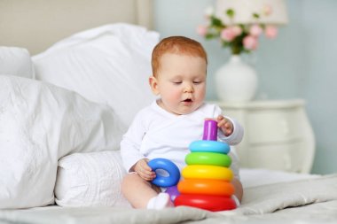 Şirin Bebek yatak beyaz güneşli yatak odasında oturup renkli gökkuşağı oyuncak piramit ile oynarken. Küçük çocuklar için oyuncaklar. Eğitici oyuncak çocuk. Erken gelişme.
