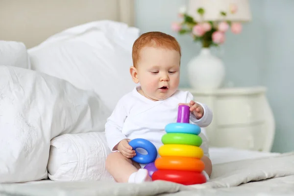 Cute baby playing with colorful rainbow toy pyramid sitting on bed in white sunny bedroom. Toys for little kids.  Child with educational toy. Early development. Royalty Free Stock Photos