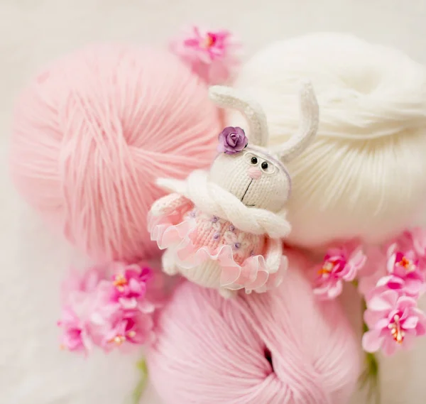Ručně pletený králík. Velikonoční zajíček v brýlích, v růžových šatech s výšivkou mezi koule příze a růžové květy — Stock fotografie