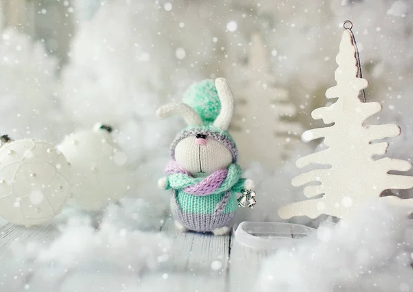 Conejo hecho a mano. Conejito de Navidad en una bufanda y sombrero con una gran bombilla entre los árboles de Navidad decorativos y bolas . Fotos De Stock
