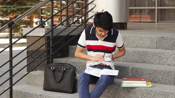 Portrait of a happy male asian student sitting on stairs and reading book — Stock Video
