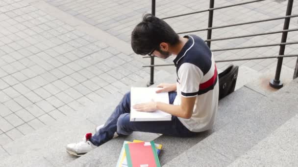 Portrait of a happy male asian student sitting on stairs and reading book — Stock Video