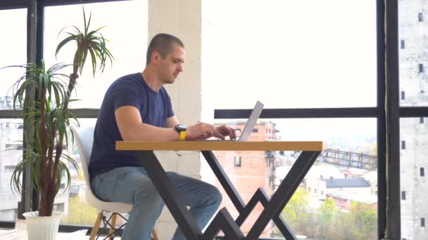 Дорослий чоловік працює на ноутбуці, сидячи в офісі з відкритим простором — стокове відео