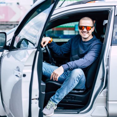 Güneş gözlüklü adam arabada oturuyor ve gülümsüyor.