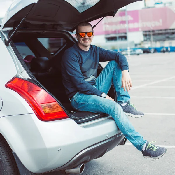 Hombre en jeans y gafas de sol sentado en el maletero abierto del coche —  Fotos de Stock