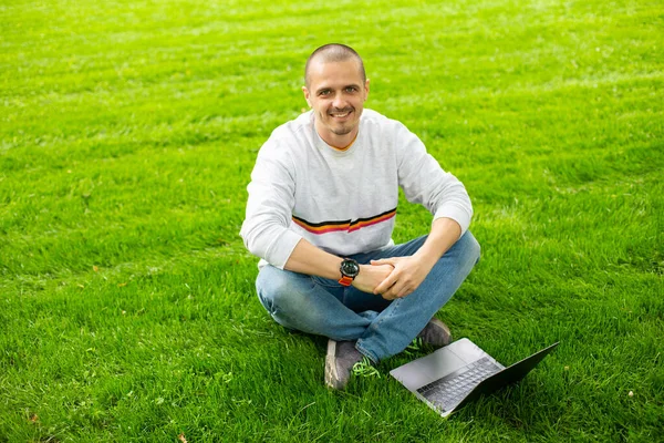 Sorrindo homem olhar na câmera e sentado no gramado verde com laptop — Fotografia de Stock