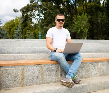 Modern şehir parkında bankta dizüstü bilgisayarı olan bir adam oturuyor.
