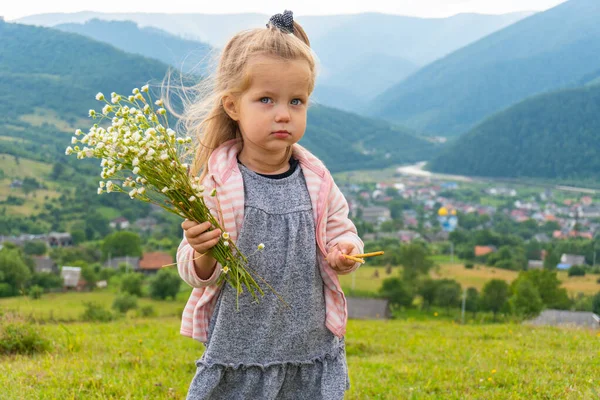 Kis komoly gyermek lány egy csokor virággal a kezében és hegyekkel mögötte. — Stock Fotó