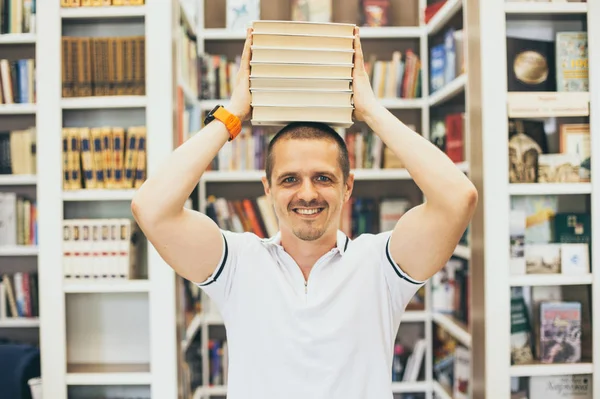 Улыбающийся мужчина с кучей книг на голове в библиотеке — стоковое фото