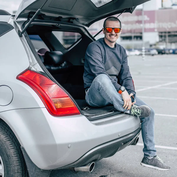 Homme souriant adulte en lunettes de soleil assis dans le coffre de sa voiture — Photo