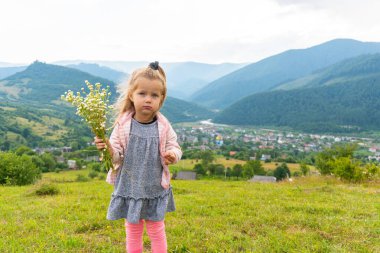 Küçük kız kameraya bakıyor ve elinde çiçekler tutuyor.