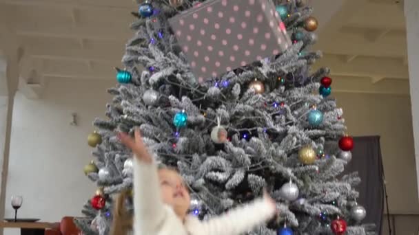 Klein schattig meisje thow tot cadeau doos met kerstboom achter — Stockvideo