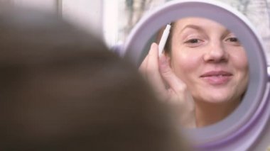 Makyaj yapan ve aynaya bakıp gülümseyen kadın.