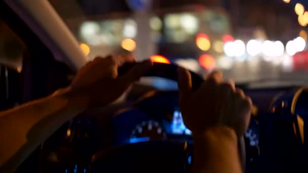 Man holding hands on steering wheel while turning left, driving car in the night city. City shining with colored lights. UHD — Stock Video