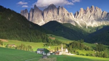 İtalya, Santa Magdalena 'daki Dolomitlerin manzarası. Dağ etekleri yeşil ormanlarla kaplıdır. Kilise ve birkaç ev ön planda. Panoramik çekim, 4k