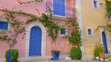 Yunanistan 'ın Kefalonia kentinde, yeşil flora ve çiçeklerle süslenmiş mavi kapı ve pencereli pastel pembe ve krem rengi evler. Sabit çekim, 4k.