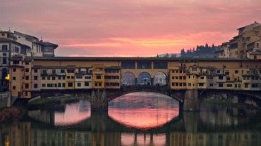 Floransa, İtalya. Sarı Ponte Vecchio Köprüsü pembe ve turuncu günbatımı gökyüzüne karşı. Gökyüzü Arno Nehri 'ne yansıyor. Döndürme atışı, Uhd